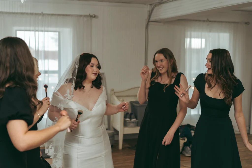 Bride laughing and getting ready with bridesmaids holding makeup brushes in a bright bridal suite.