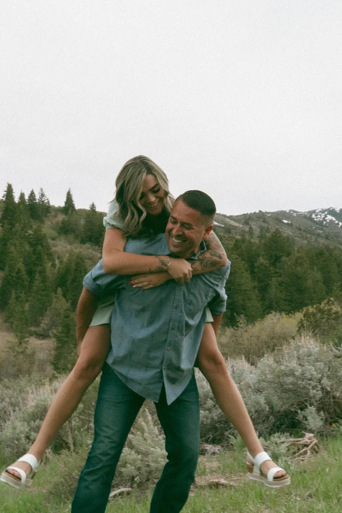 Man gives woman a piggyback ride on a grassy mountainside, both laughing together.