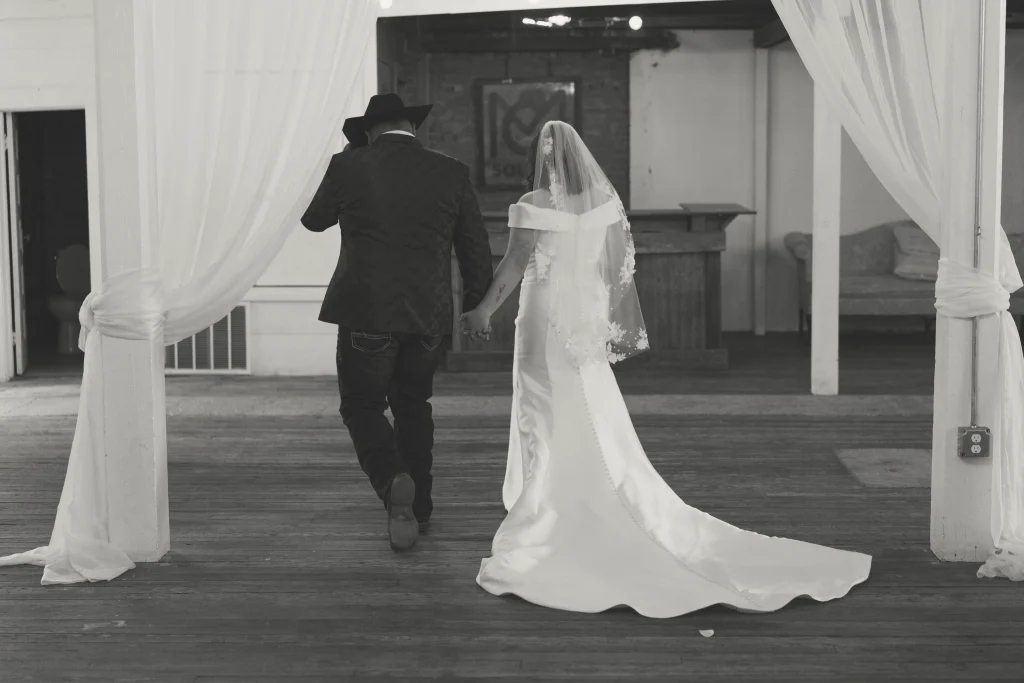 Bride and groom holding hands while walking into their venue, captured from behind in black and white.