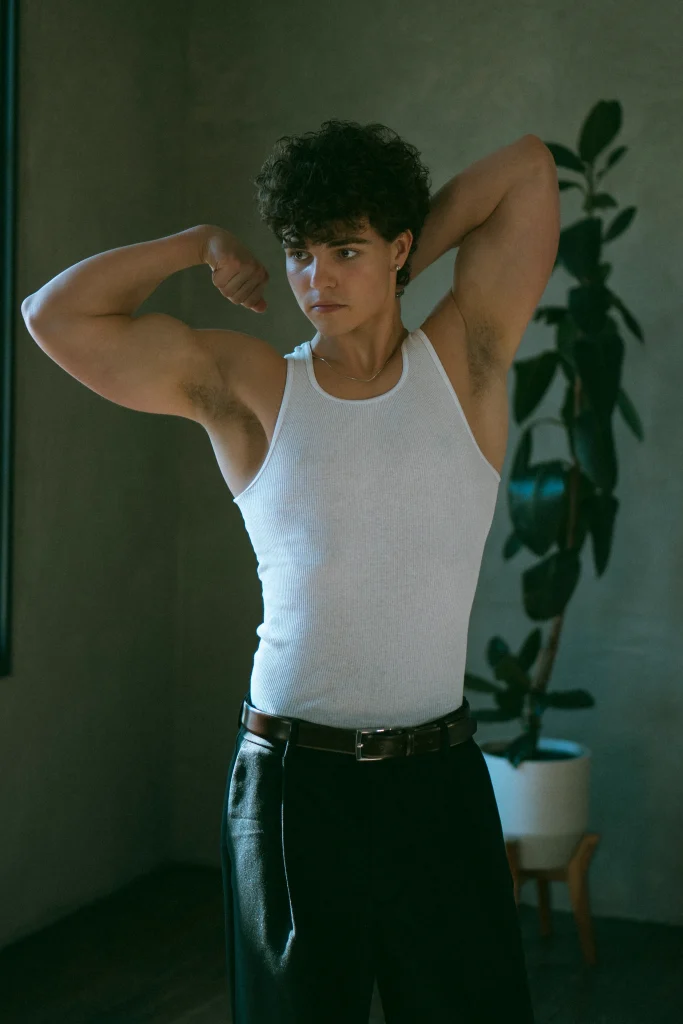 Young man flexing his arms in a white tank top, posing indoors next to a plant.