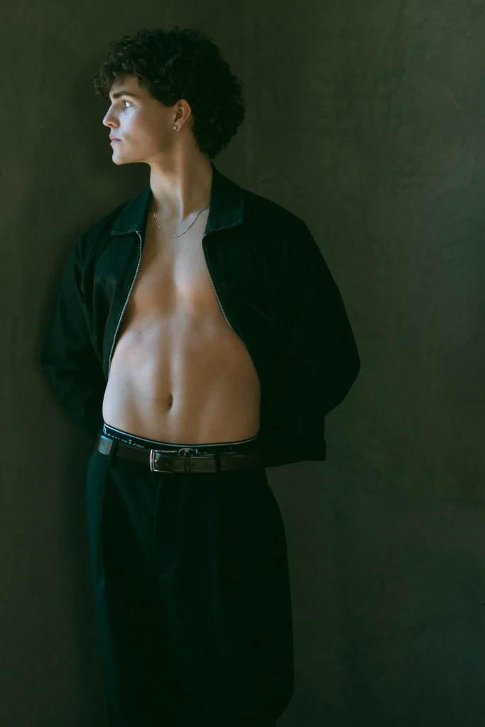 Young man standing shirtless with an open jacket, looking to the side in soft natural light.