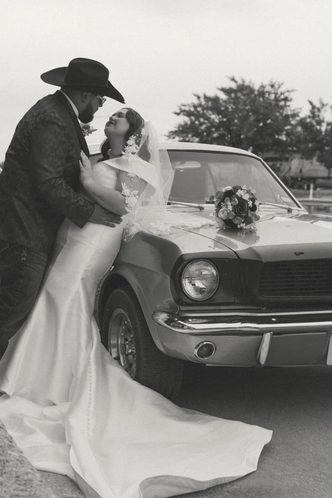 Bride leaning against classic car while groom holds her close in a romantic black and white moment.