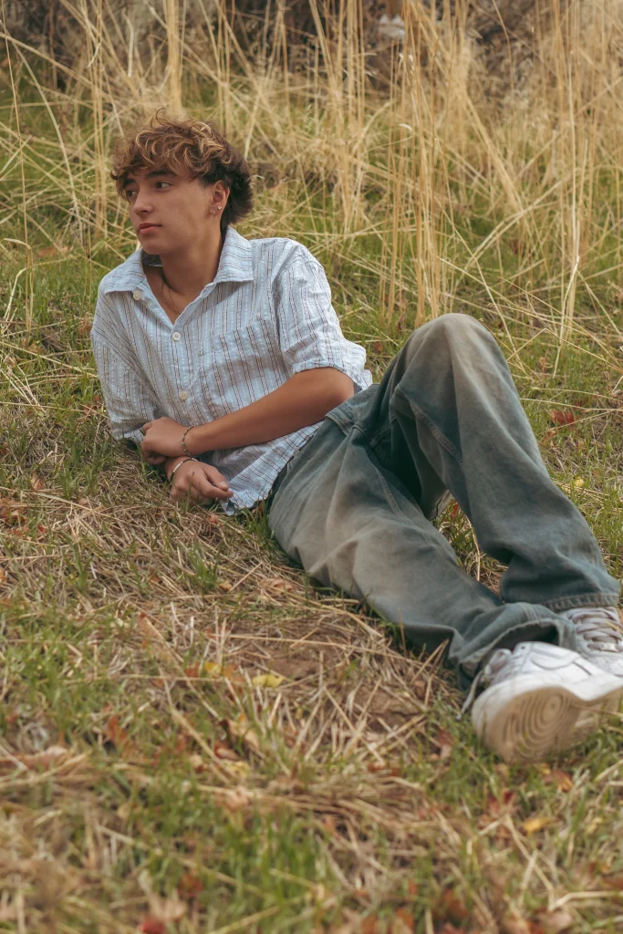 Teen boy lying in tall dry grass, wearing a light striped shirt and looking to the side.