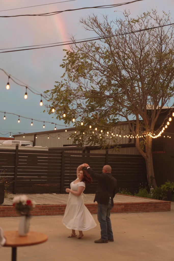 Bride and groom dancing outdoors under string lights with a faint rainbow in the sky.