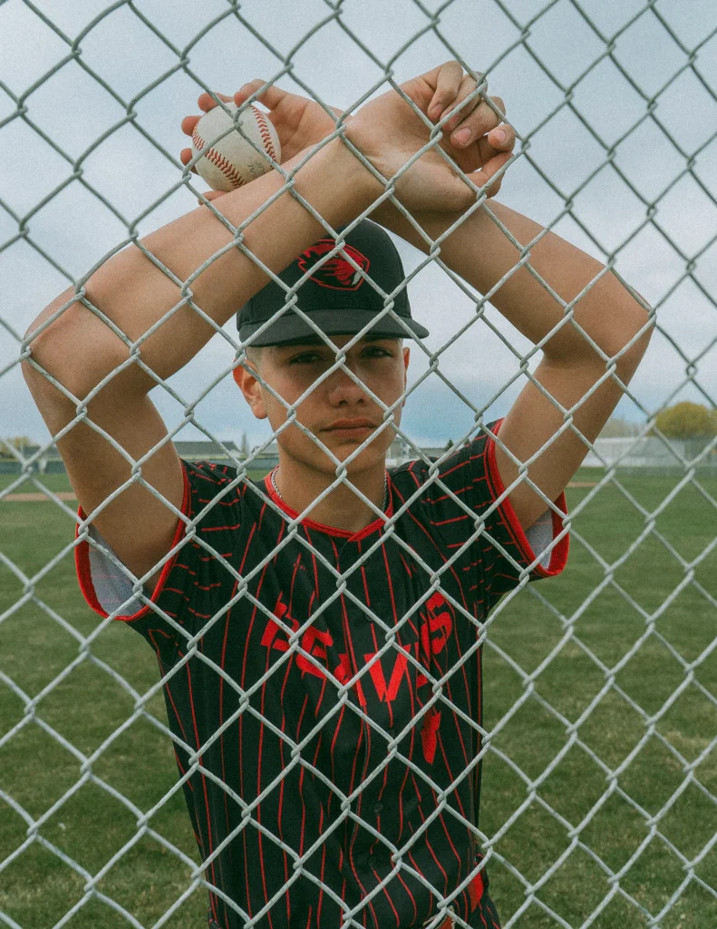 Baseball player standing behind a chain-link fence holding a ball above his head.