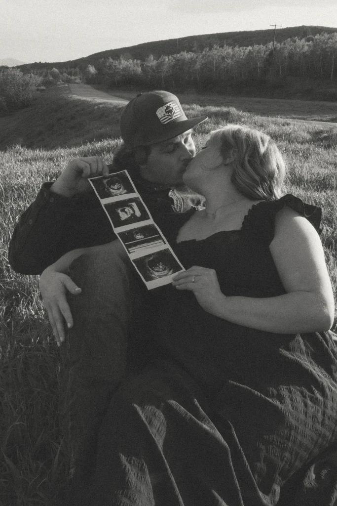 Black-and-white image of a couple kissing while holding ultrasound photos in a grassy field.