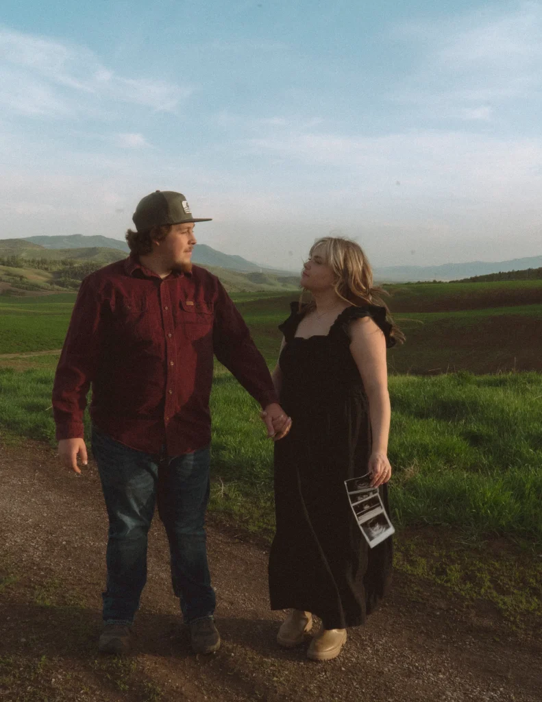A pregnant woman sits on a blanket in a grassy field with her partner, holding ultrasound photos as they walk together at sunset.