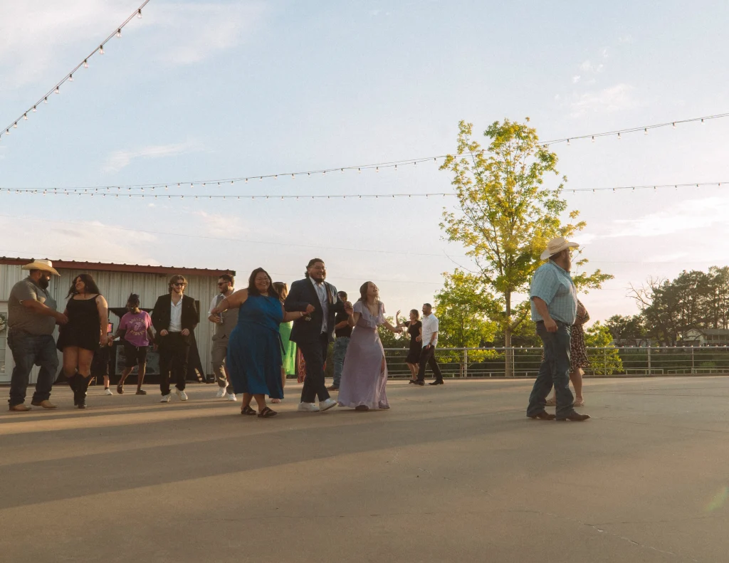 Wedding guests and the couple dancing outdoors at sunset under string lights.