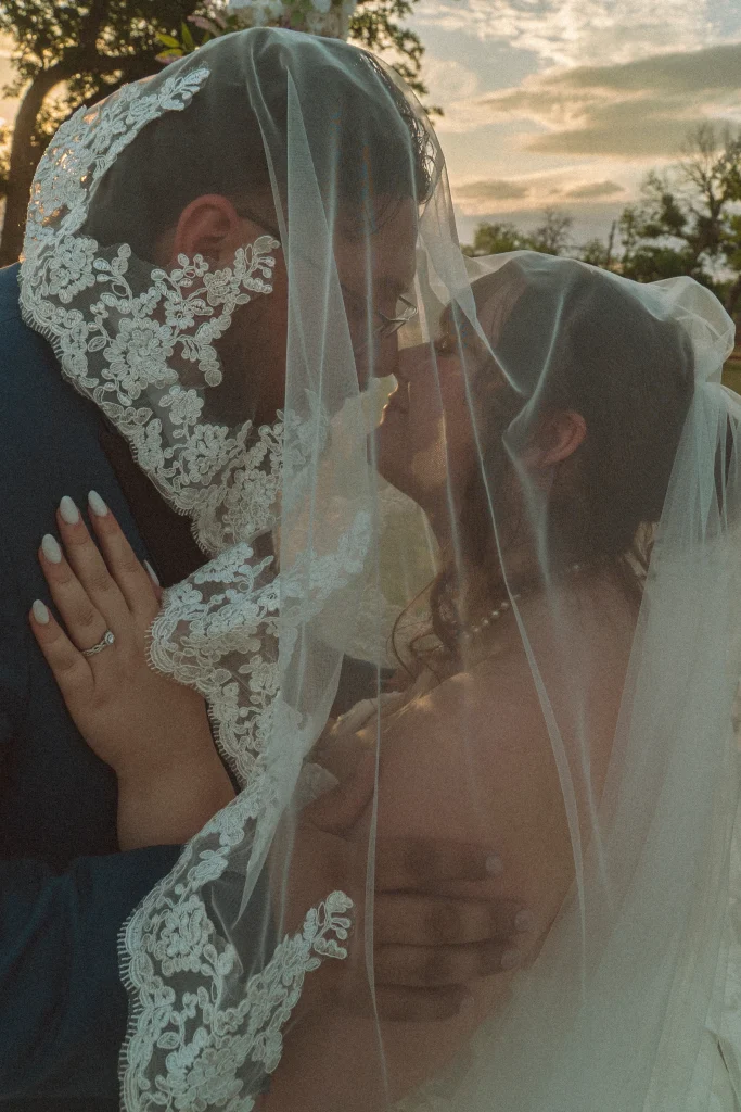 Bride and groom sharing a kiss beneath the bride’s lace veil during golden hour.