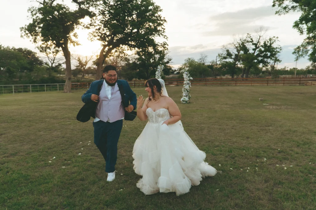 Bride and groom laughing and running together across a grassy field after their ceremony.