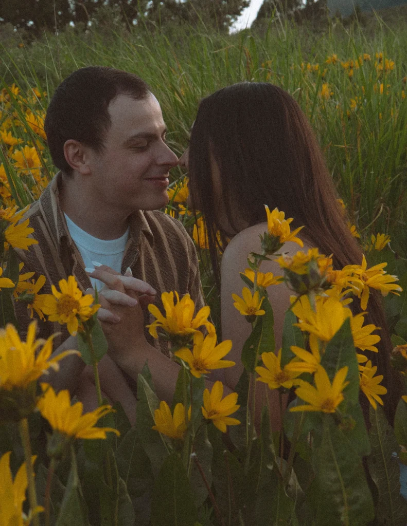 Couple touches noses while holding hands in a field of blooming yellow flowers.