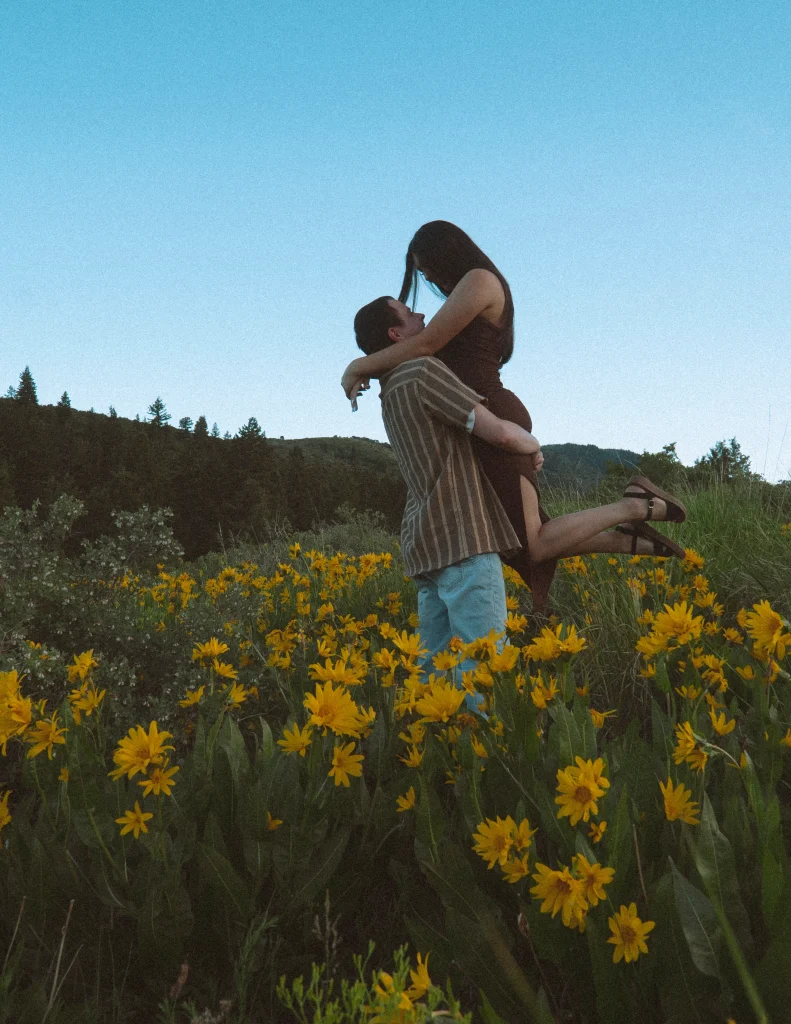 Man lifts woman in a field of yellow wildflowers with mountains in the background.