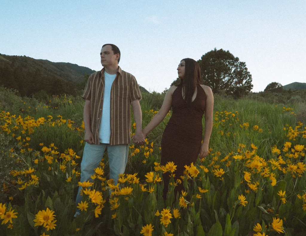 Couple stands hand-in-hand surrounded by tall yellow flowers and mountain scenery.