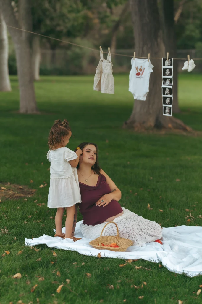 A pregnant woman sits on a blanket with her young daughter standing beside her, with baby clothes and ultrasound photos hanging on a clothesline behind them.