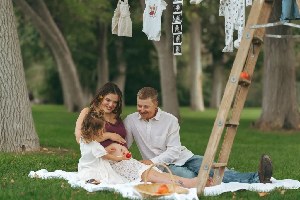 A family sits together on a picnic blanket in a park, smiling as their young daughter holds fruit near the mother's pregnant belly; baby items hang on a clothesline above.