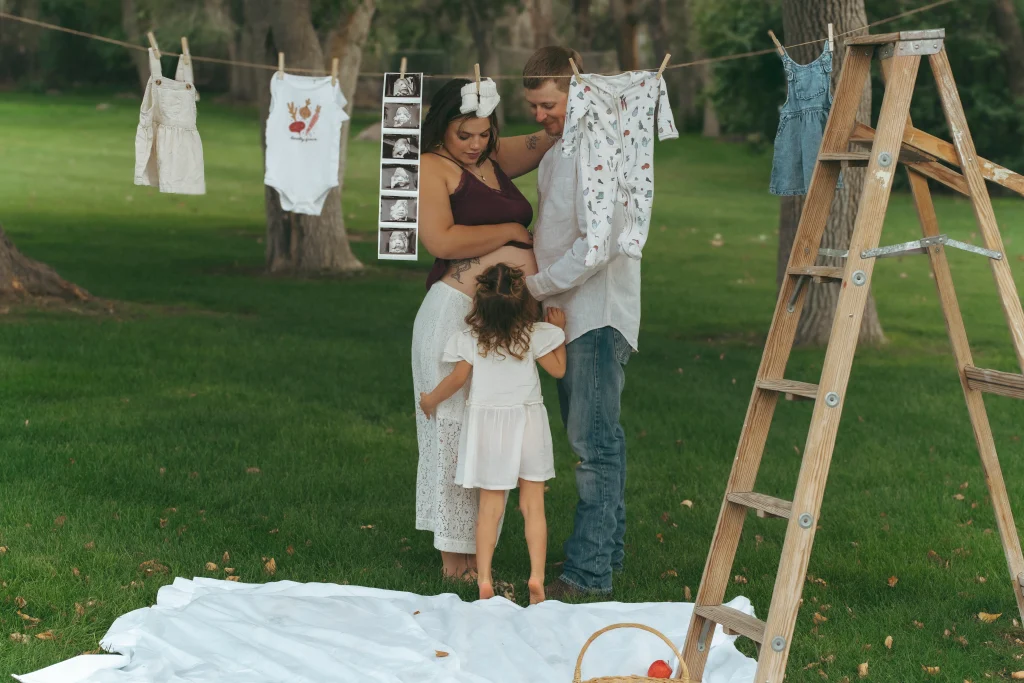 A family of three stands beneath a clothesline of baby clothes and ultrasound photos, with the child touching the pregnant mother’s belly.