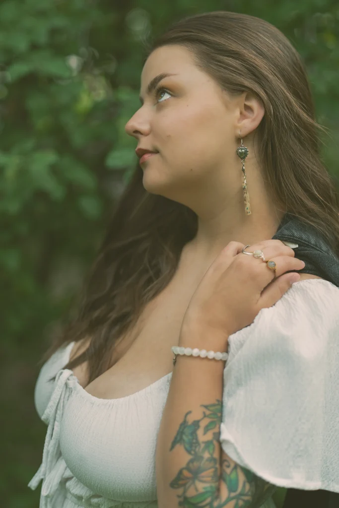 Woman’s reflection in a handheld mirror outdoors, resting her chin on her hand and wearing multiple rings, a bracelet, and long earrings.