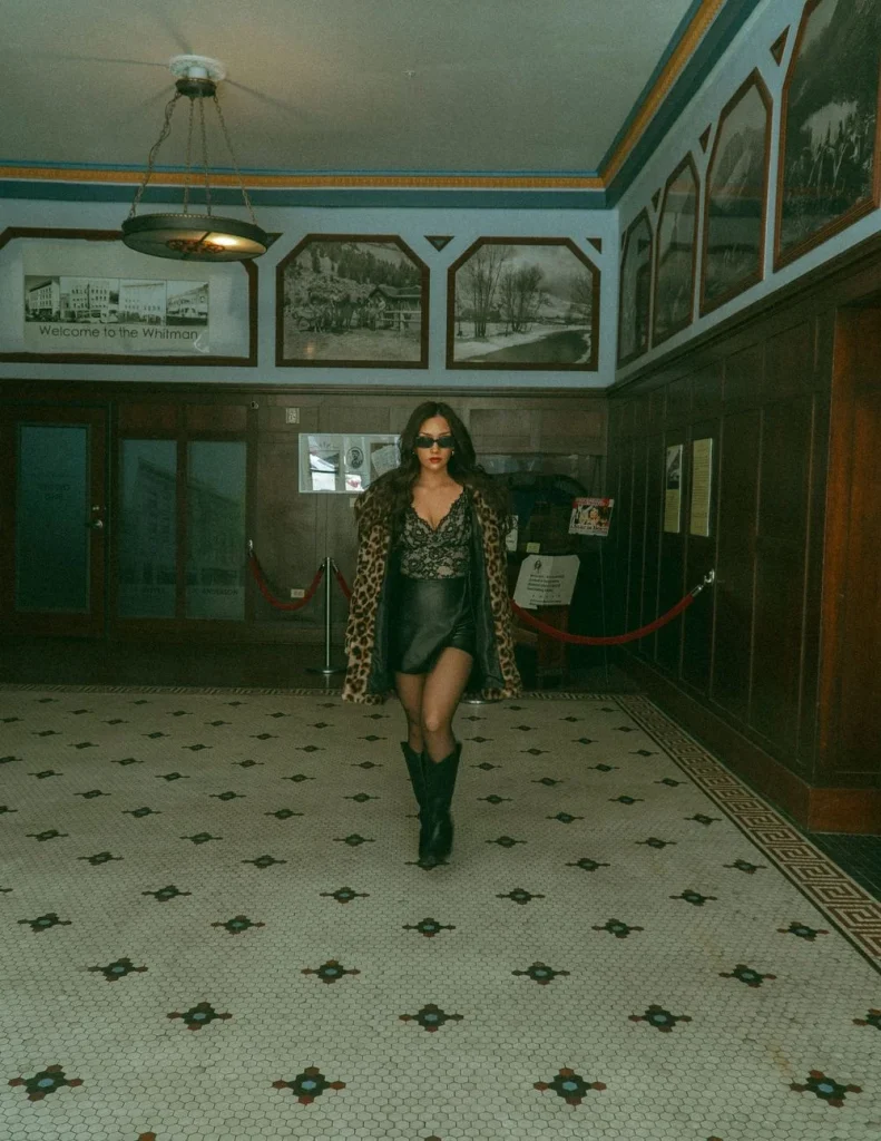 Woman walking confidently across a tiled lobby in a historic building, wearing a leopard coat, lace top, and boots.