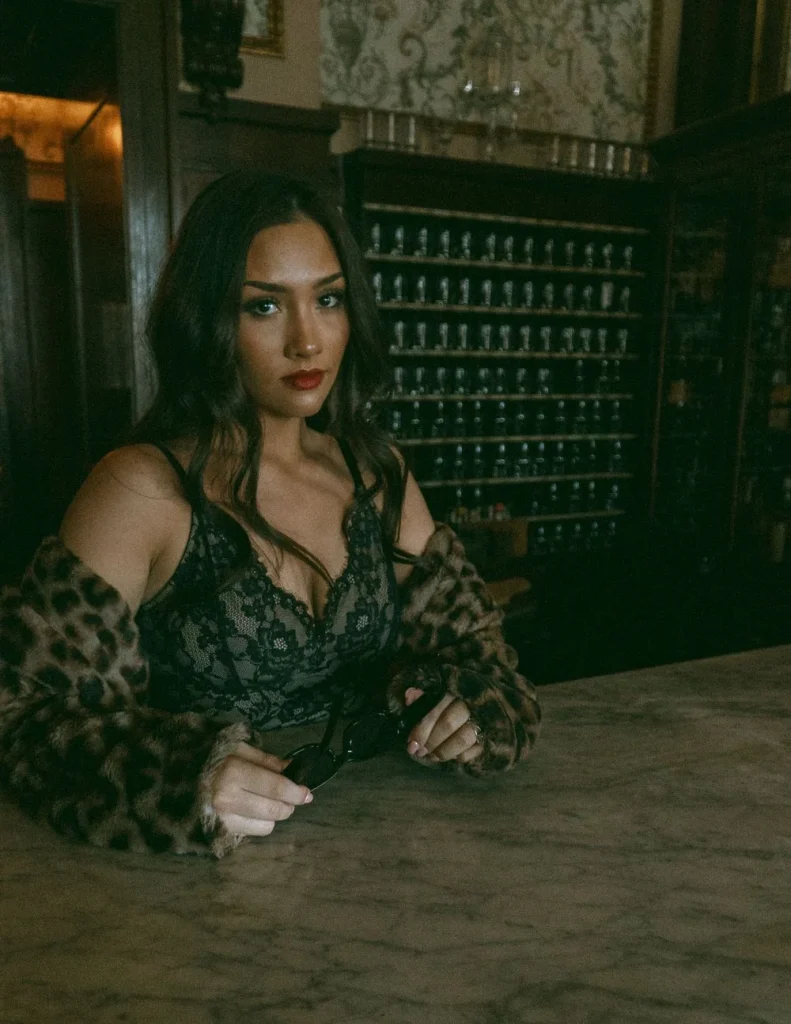 Woman leaning on a marble counter in a dimly lit room, wearing a lace top and holding sunglasses.