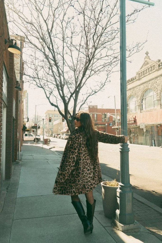 Woman standing outdoors on a sunlit sidewalk, holding a lamp post while wearing a leopard coat and boots.