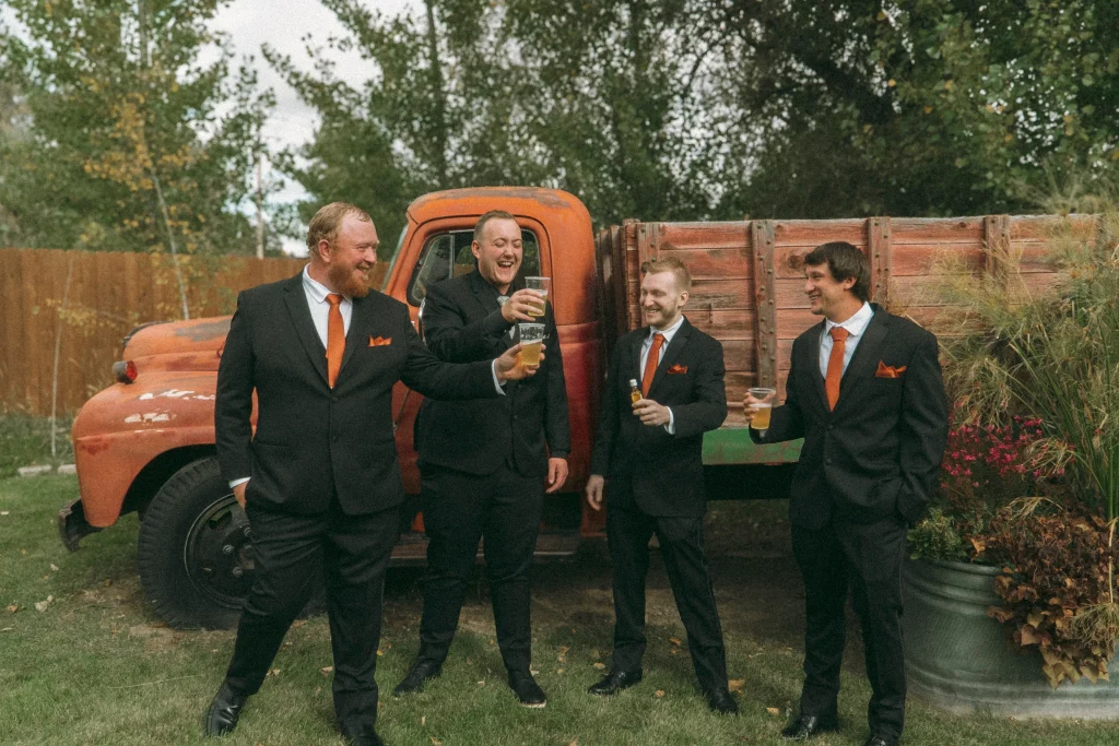 Groomsmen in black suits with orange ties sharing drinks and laughing in front of a vintage red truck.