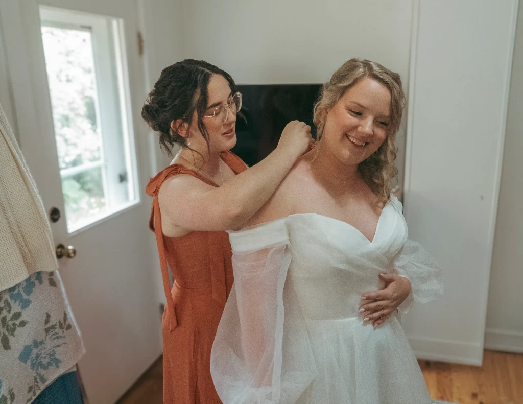 Bridesmaid helping the bride adjust the back of her dress as the bride smiles during getting-ready moments.