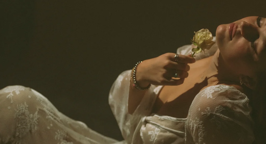 Close-up of a woman reclining with a white rose held to her chest, her lace dress illuminated in soft shadows.