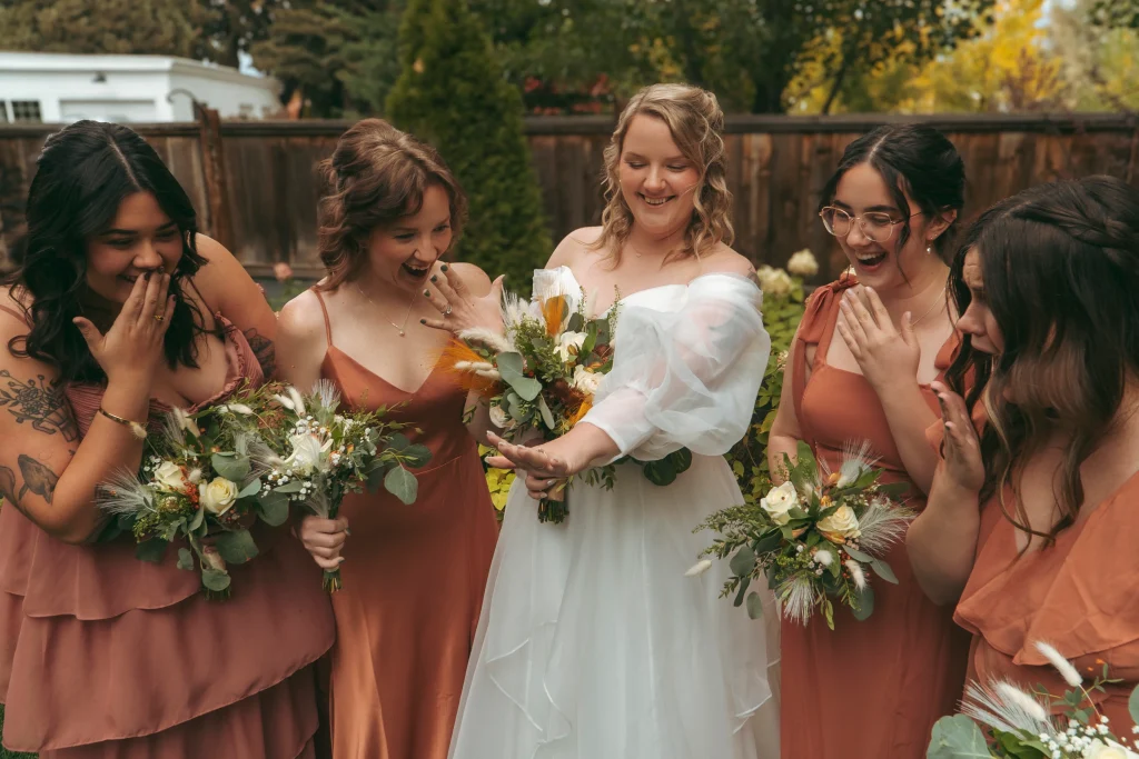 Bride showing her wedding ring to bridesmaids who react with excitement while holding bouquets.
