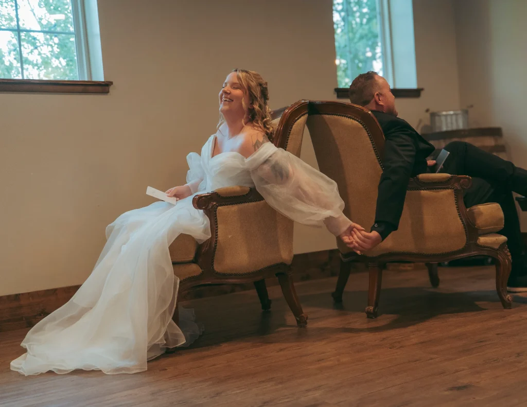 Bride and groom sitting back-to-back in chairs, holding hands and smiling during a pre-ceremony moment.