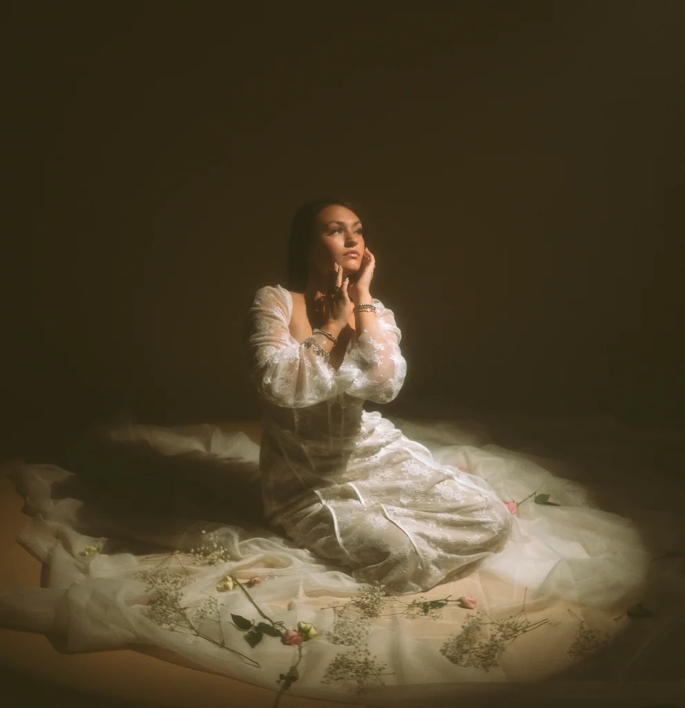 Woman sitting in a circular spotlight on layers of tulle and scattered flowers, wearing a flowing white lace gown.