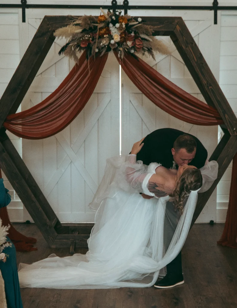 Groom dipping the bride for a kiss beneath a floral hexagon arch indoors.