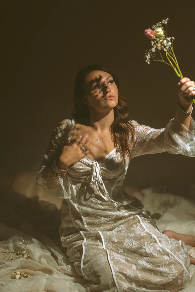 Woman in a white lace dress holding a small bouquet up toward the light, shadows from the flowers crossing her face.