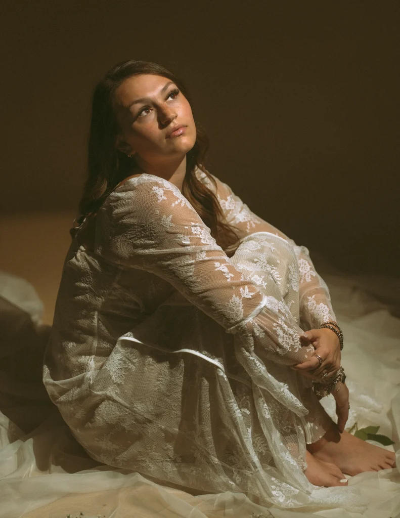 Woman seated with her knees drawn in, wearing a long white lace dress, looking upward in warm, dramatic lighting.