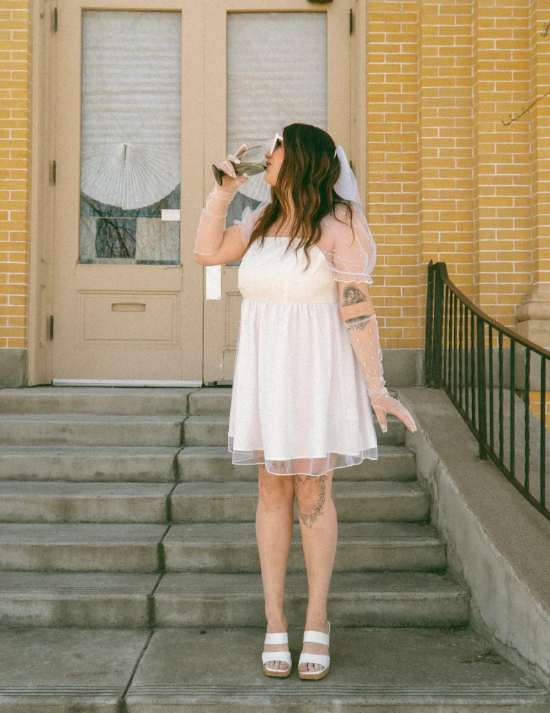 Woman in a short white dress and veil standing on stone steps, sipping from a wine glass in front of a yellow brick building.