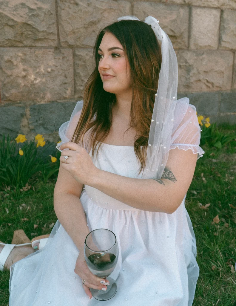 Woman sitting on the grass in a white dress and veil, holding a wine glass and looking off to the side.