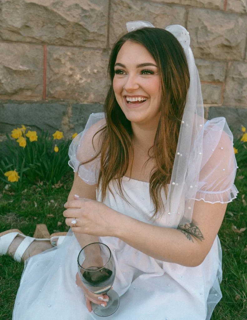 Woman in a white dress and veil sitting on the grass, laughing while holding a wine glass, with flowers blooming behind her.