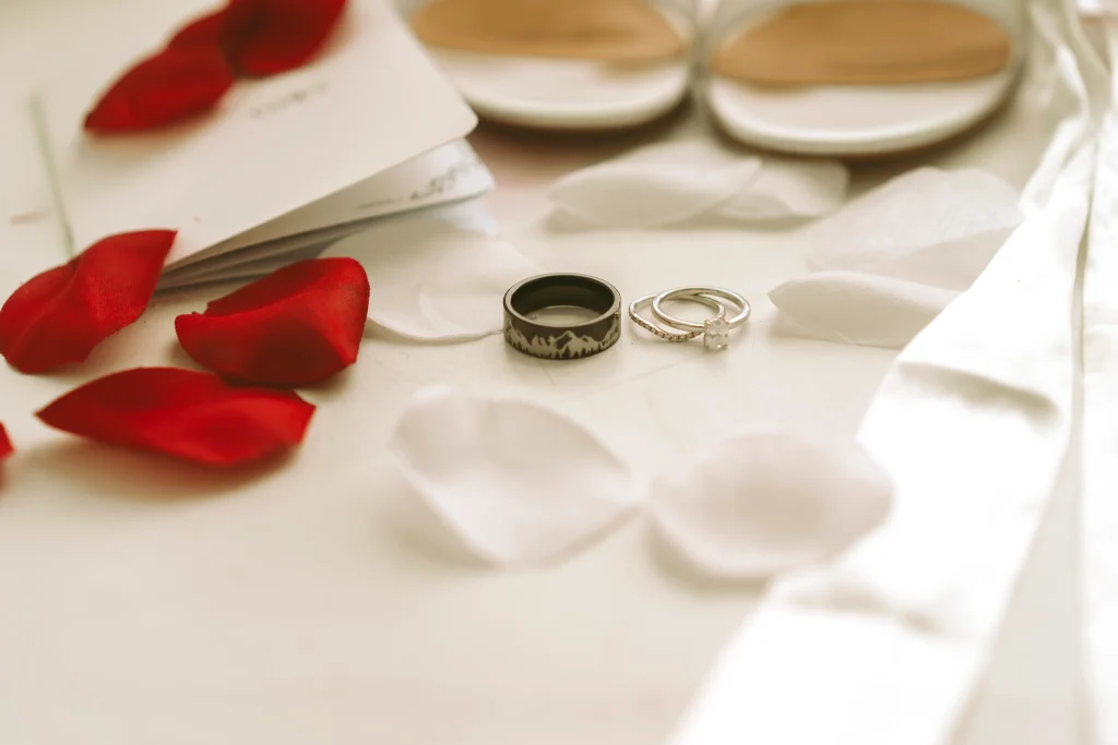 Wedding rings arranged with red and white rose petals, shoes, and vow books on a table.