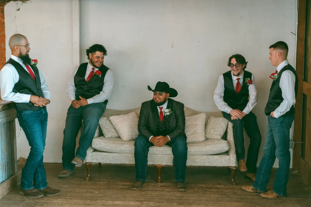 Groom and groomsmen in vests and ties laughing together while gathered around a vintage sofa.