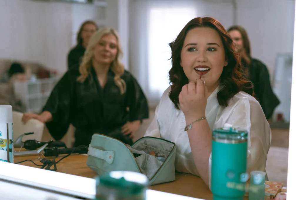 Bride applying lipstick in a mirror while bridesmaids watch during getting-ready moments.