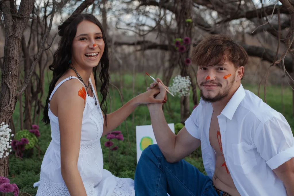 Couple sits in a garden painting each other with bright colors, smiling at the camera.
