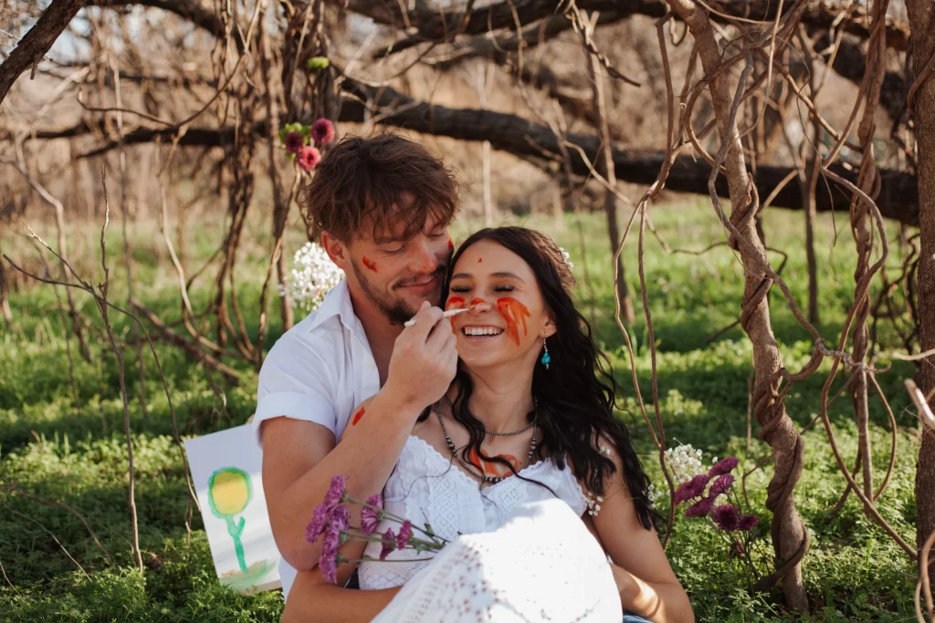 Couple laughing as the man paints the woman's face with orange paint while sitting together in a wooded area.