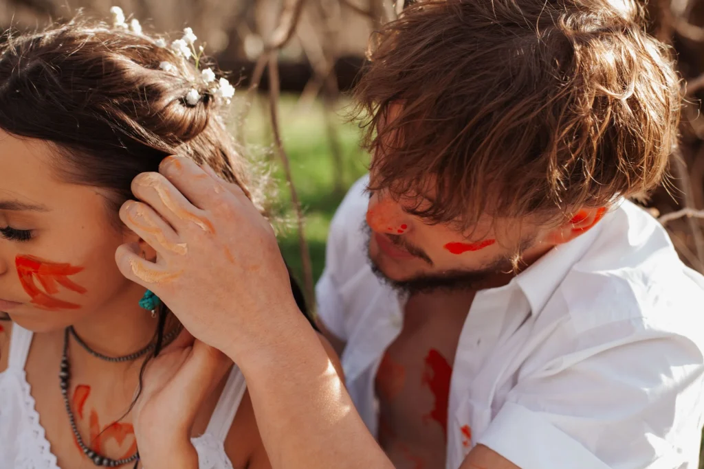 Man gently tucking flowers into the woman’s hair while both have colorful paint on their faces.