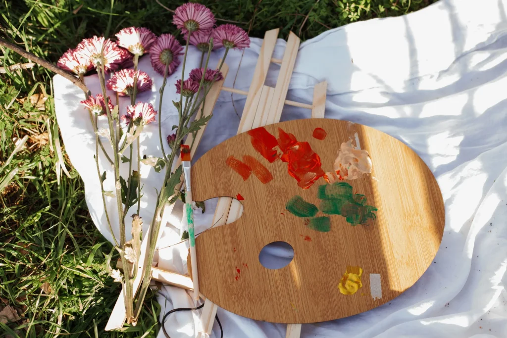 Close-up of a paint palette, brush, and purple flowers resting on a white cloth outdoors.