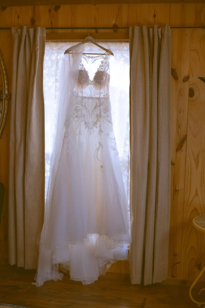 Wedding dress hanging in a wooden room, backlit by window light between curtain panels.