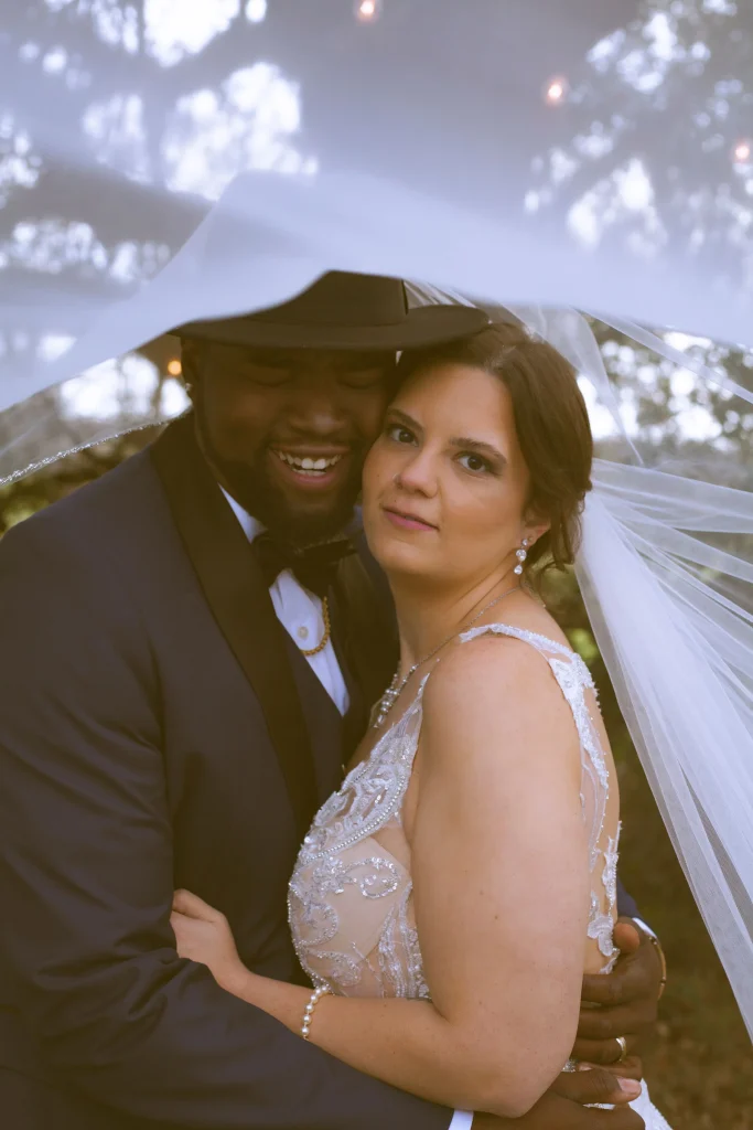 Bride and groom embracing under the bride’s veil, with the groom smiling and the bride looking toward the camera.