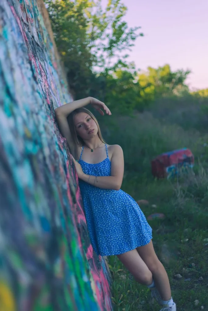 Young woman in a blue dress leaning against a colorful graffiti wall with her arm raised.