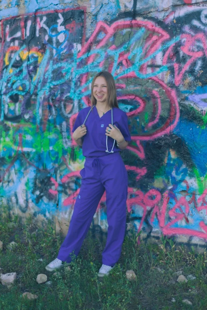 Young woman in purple scrubs smiling in front of a vibrant graffiti wall.