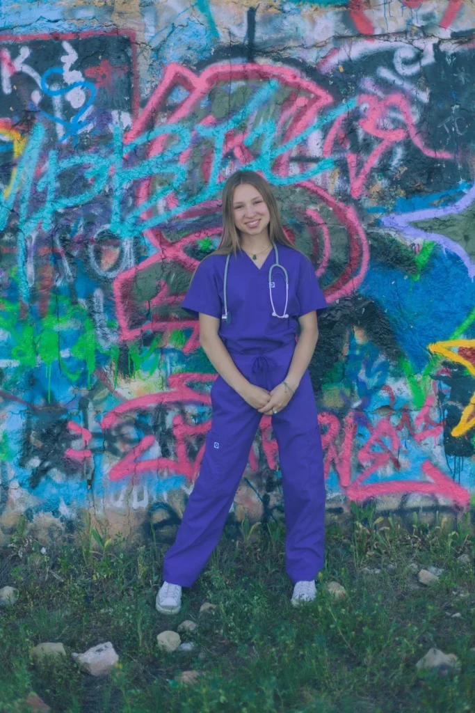 Young woman in purple scrubs holding a stethoscope and standing before a graffiti-covered wall.