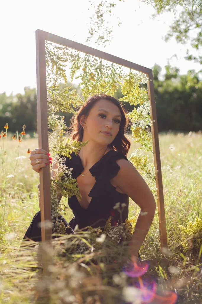Woman holding a flower-decorated frame in a sunlit field, looking off to the side.