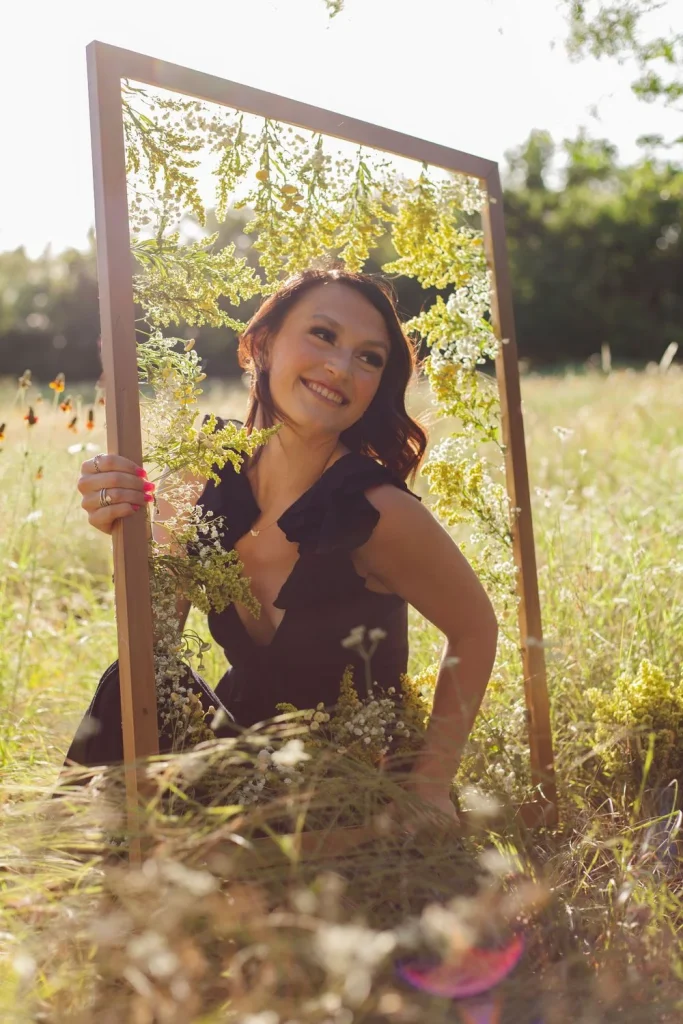Woman smiling inside a flower-decorated frame in a bright meadow.